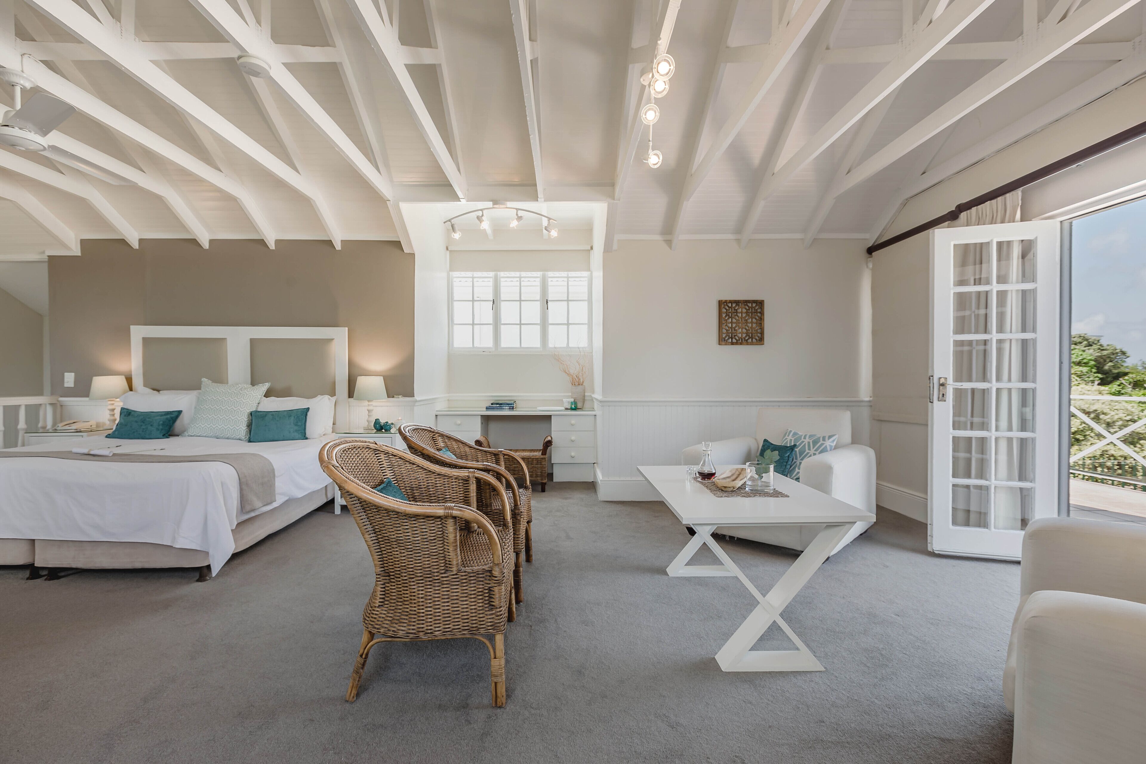 A spacious bedroom with a large bed, a desk, wicker chairs, and a view through a sliding glass door.