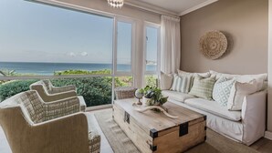 Un salon spacieux avec une grande fenêtre donnant sur l’océan, un canapé blanc et une table basse en bois.