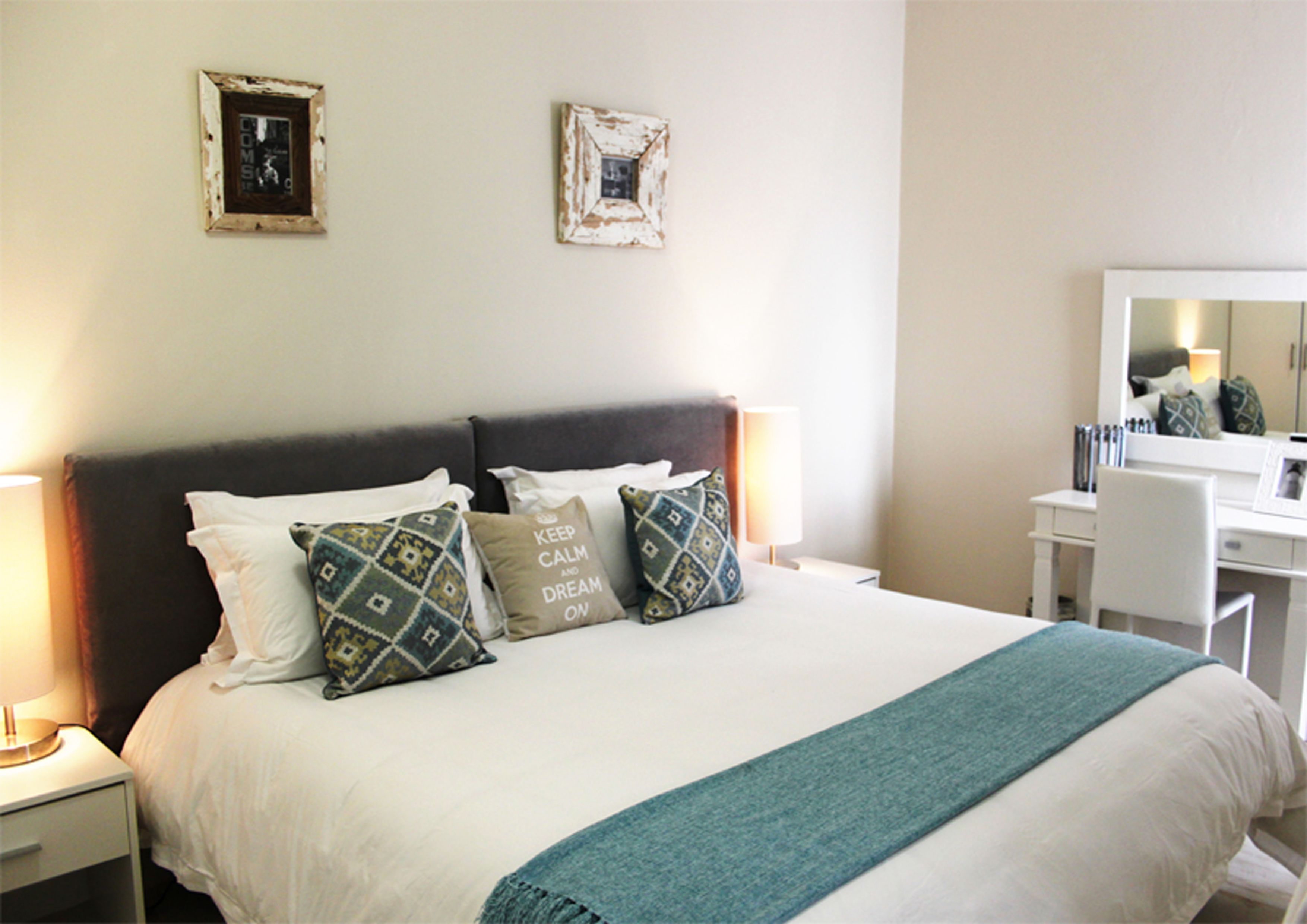 A neatly made bed with patterned pillows, a bedside lamp, and a desk with a chair and mirror.
