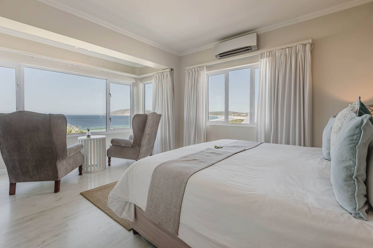 Une chambre à coucher avec un grand lit, deux fauteuils, une petite table ronde et une vue sur la mer grâce à de grandes fenêtres.