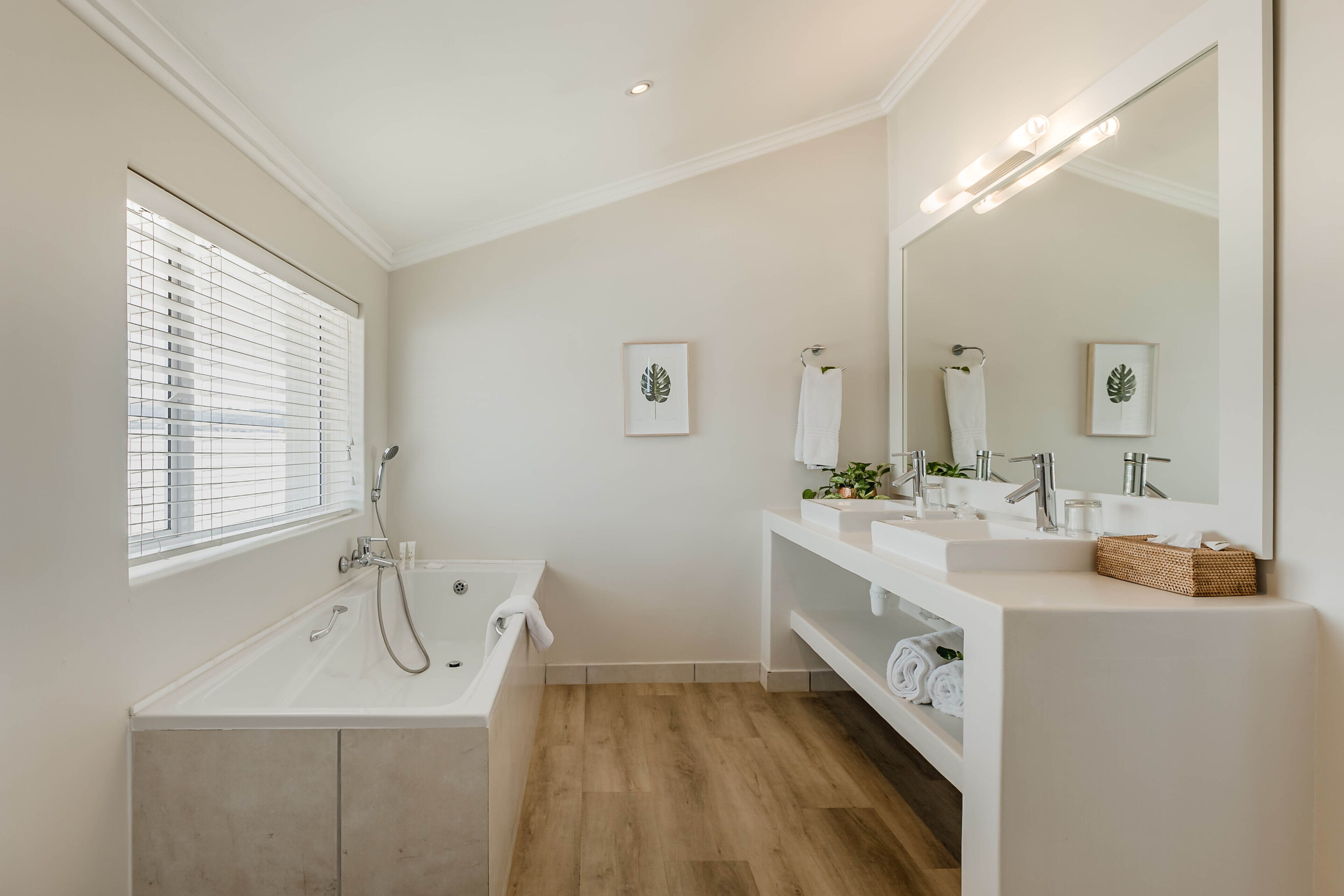 A modern bathroom with a bathtub, two sinks, a large mirror, and a window with blinds.
