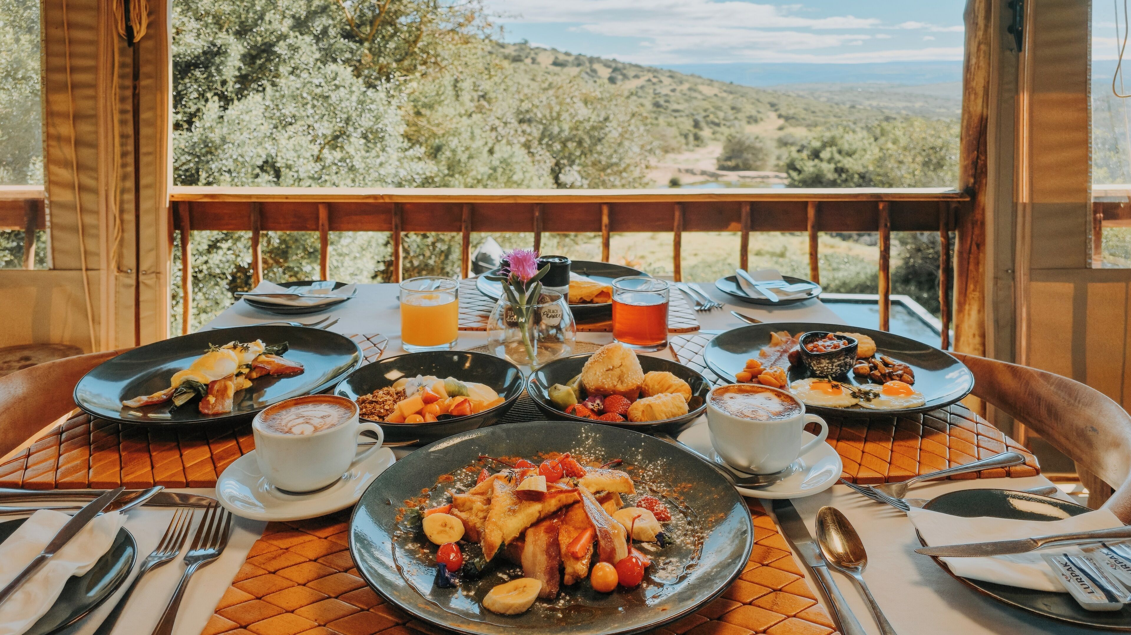 A breakfast table with various dishes, drinks, and utensils, set against a scenic outdoor view.