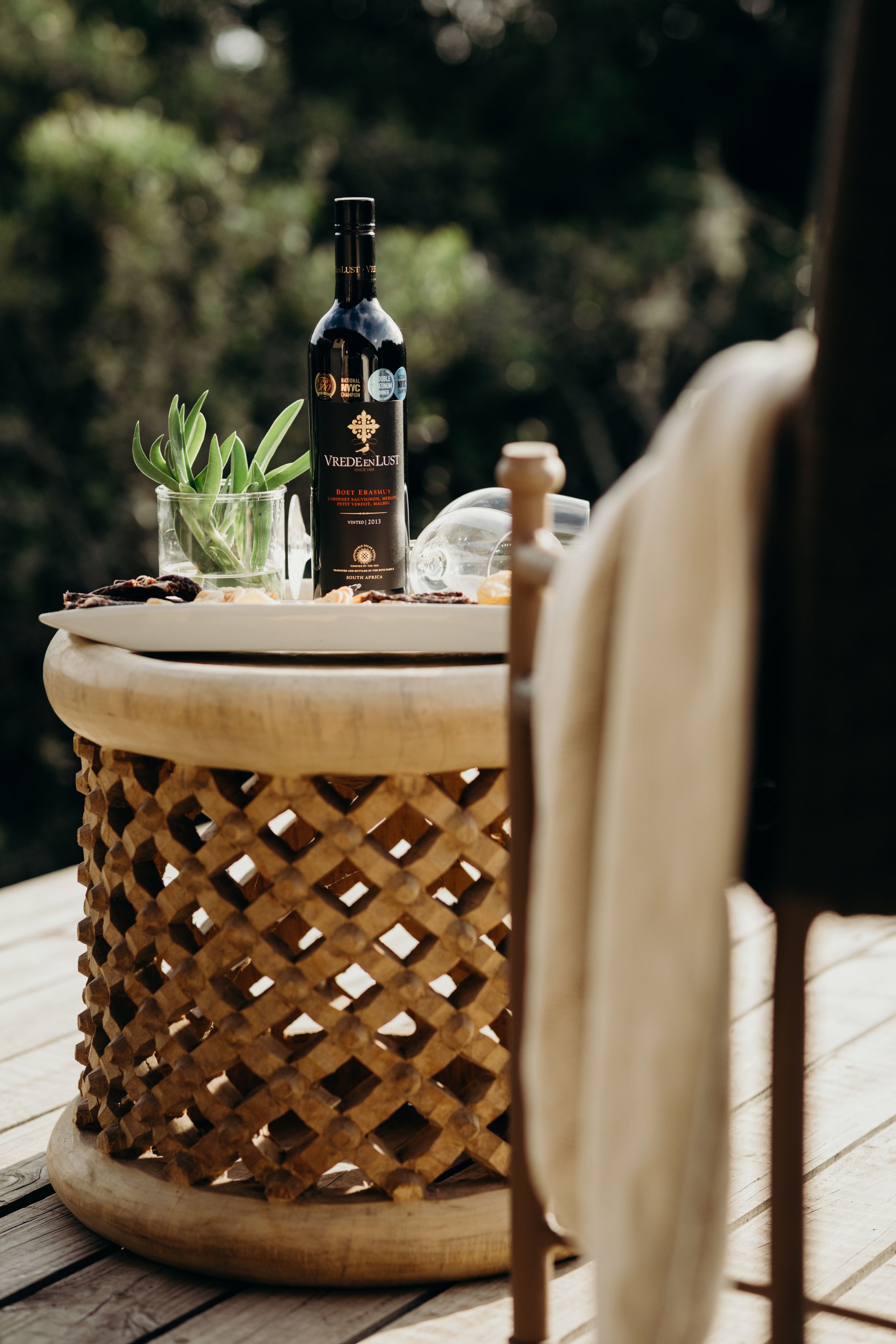 Uma garrafa de vinho Vredeblust sobre uma mesa de madeira redonda, com uma cesta trançada, um copo de água e uma pequena planta.