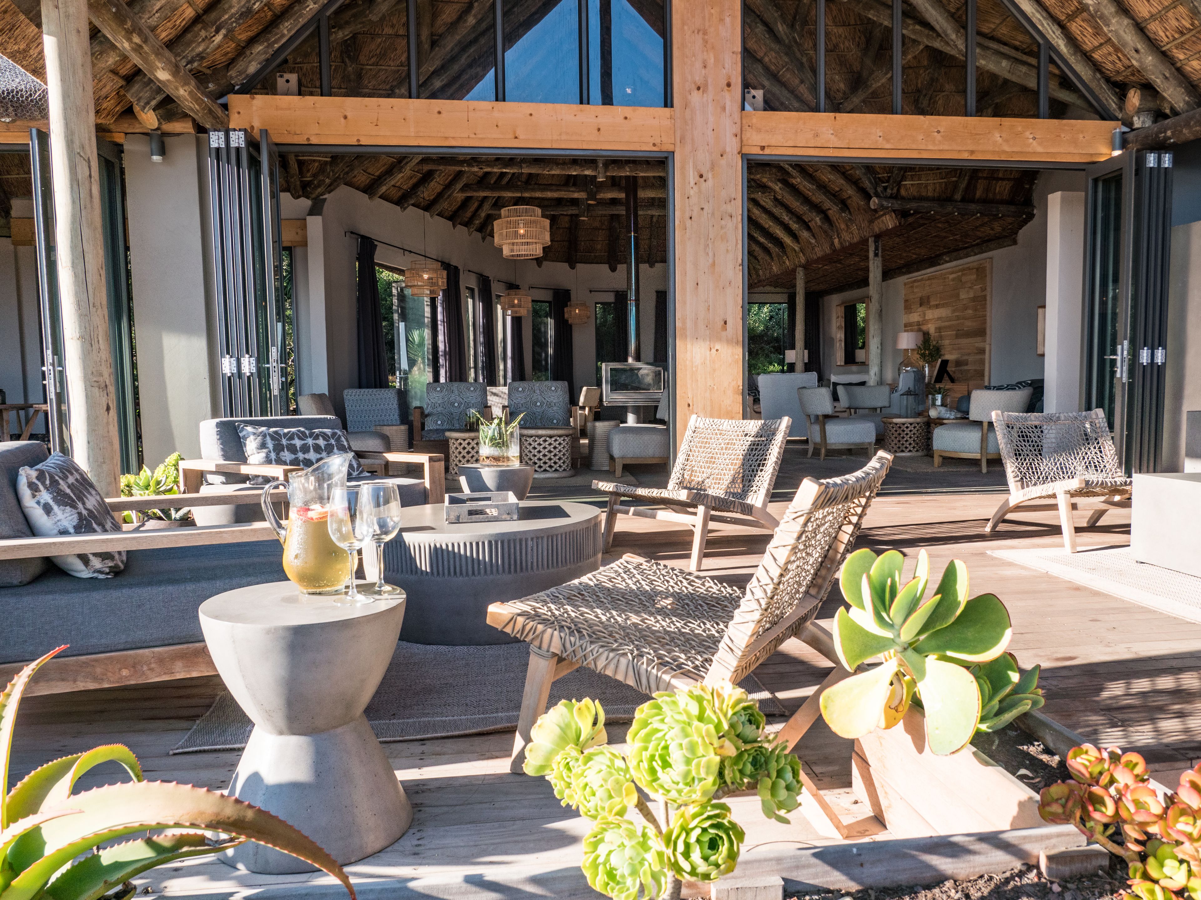 Un espace de détente extérieur abrité, doté de différents types de sièges, notamment des canapés et des fauteuils, ainsi qu’une table centrale sur laquelle sont disposées des boissons et une carafe.