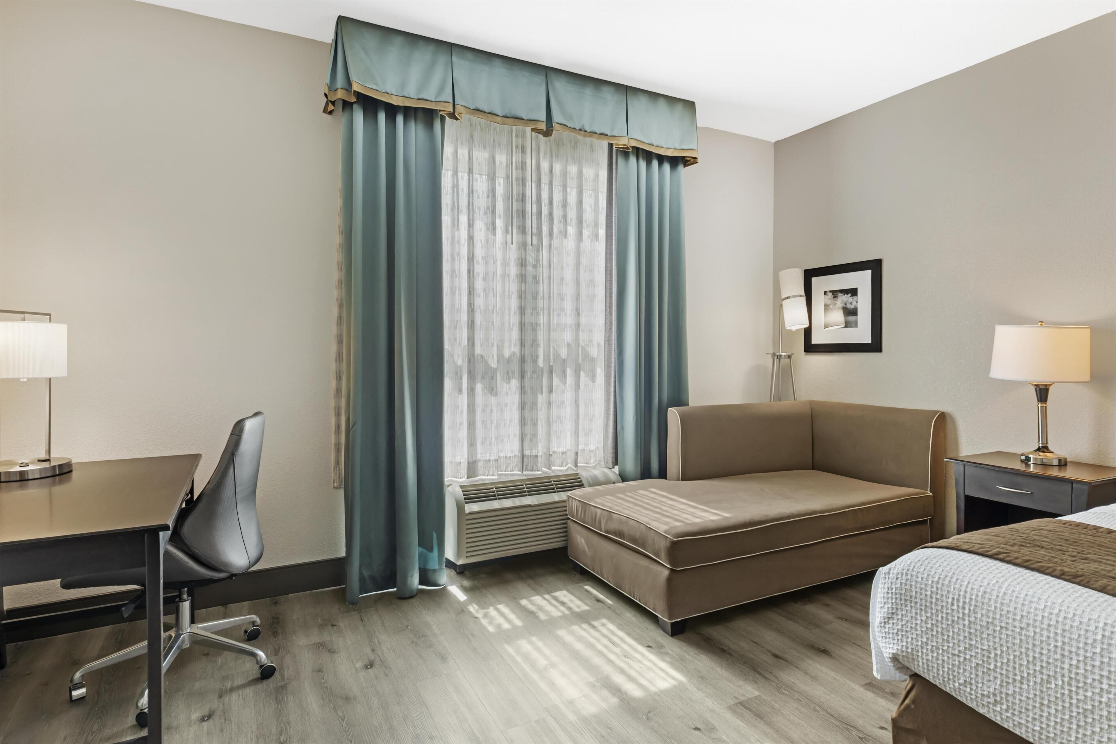 A hotel room with a desk, office chair, sofa, bed, bedside table, lamp, and a picture on the wall.