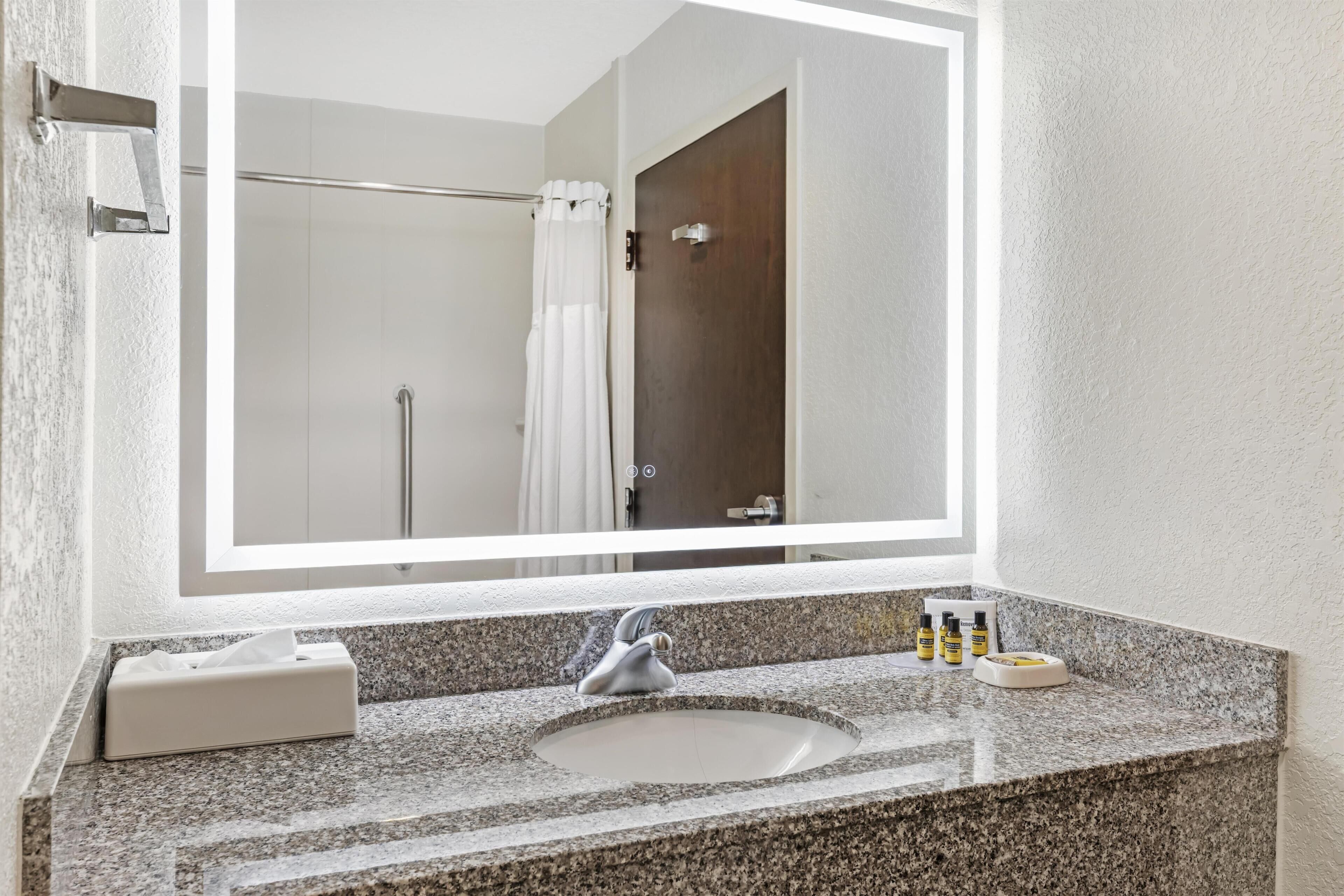 Baño con encimera de granito, lavamanos, espejo, dispensador de toallas y cortina de ducha.