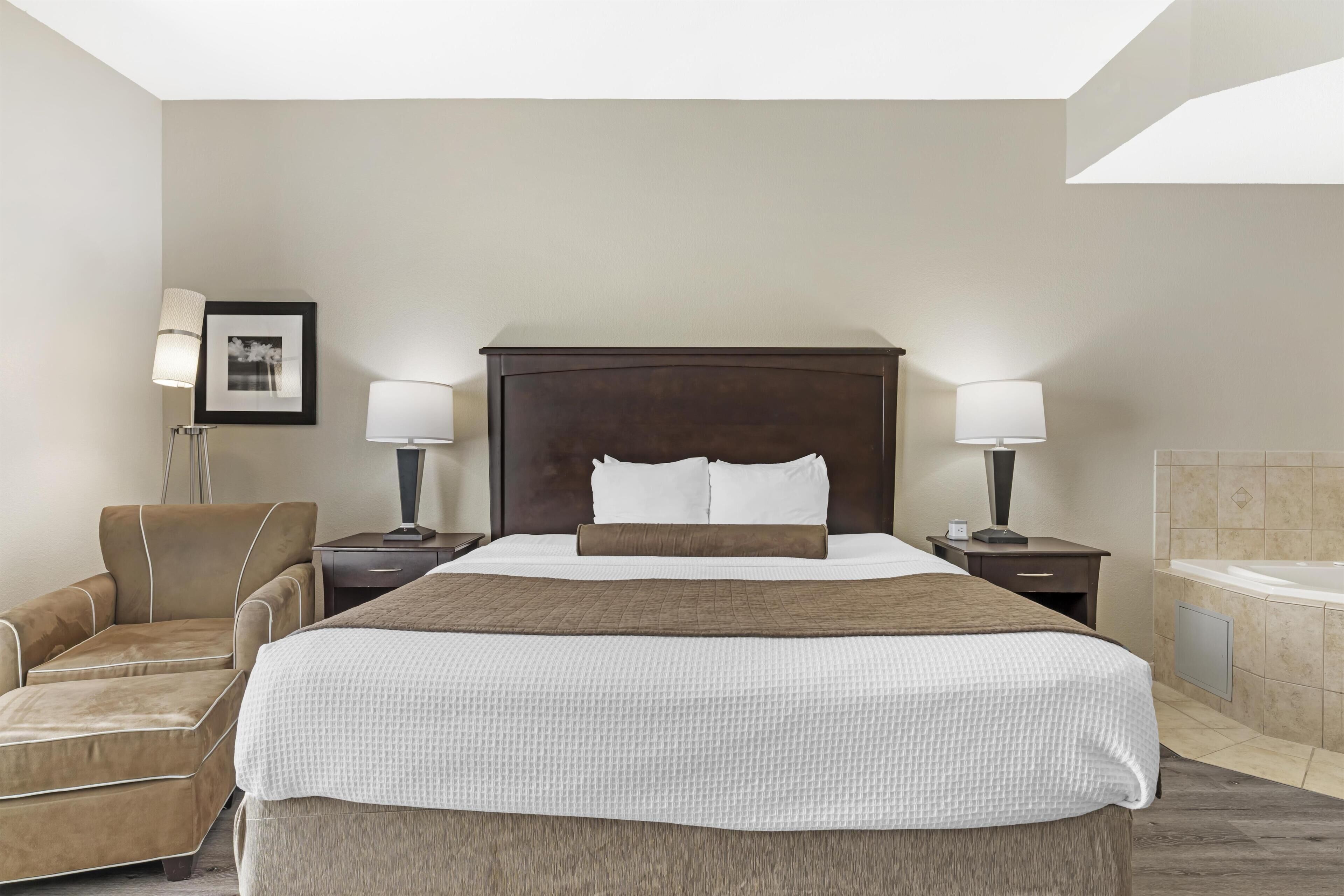 Habitación de hotel con una cama grande, una silla, una mesita de noche, una lámpara y un baño con bañera.