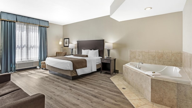A hotel room with a bed, bedside tables, a desk, a chair, a large window with curtains, a bathtub, and a ceiling light.