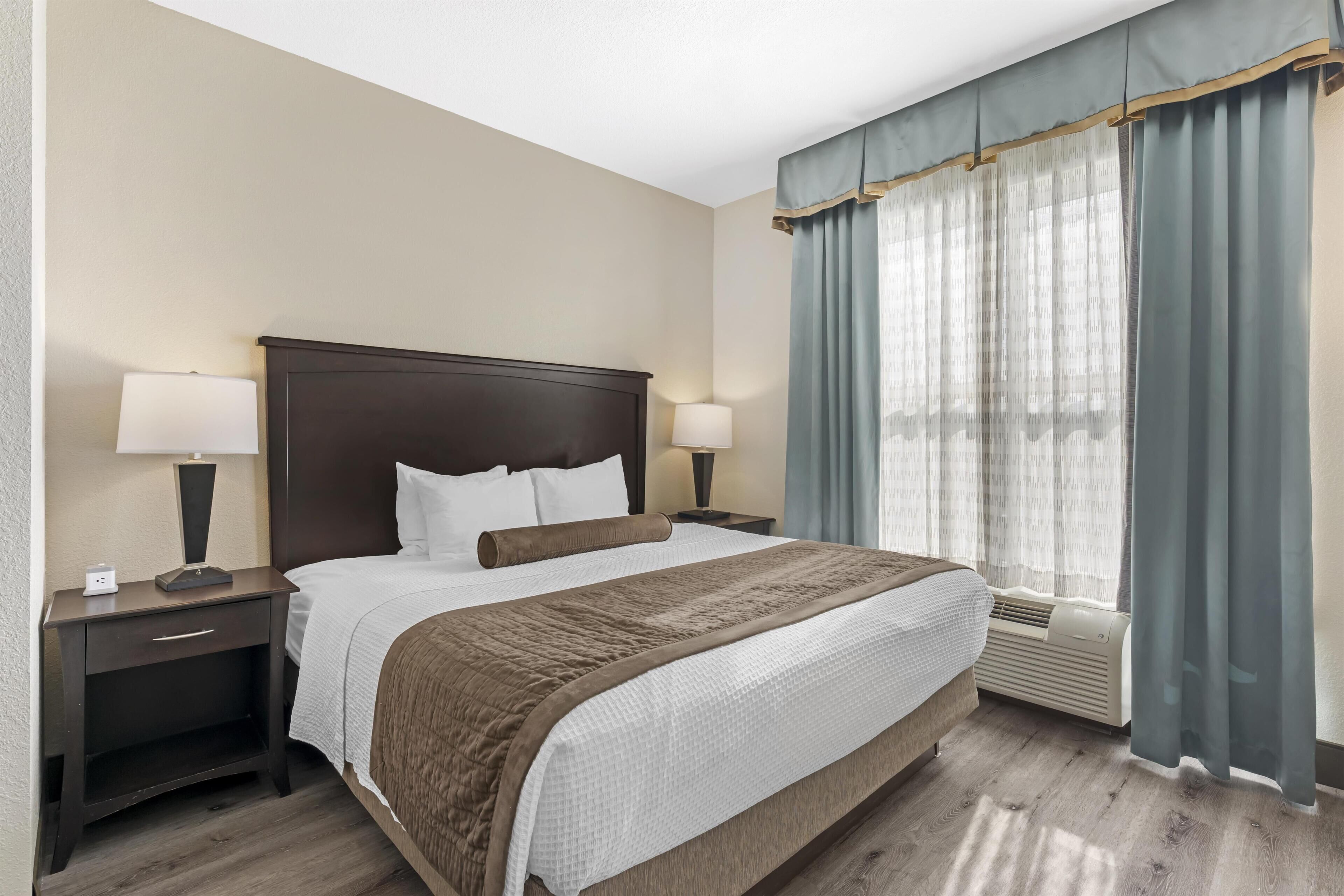 A hotel room with a large bed, two bedside tables, a nightstand, a window with curtains, and a wall-mounted air conditioning unit.