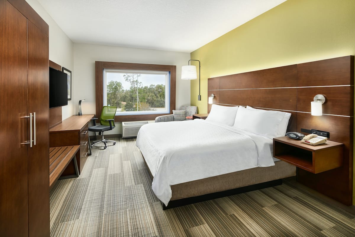 A hotel room with a large bed, a desk, a chair, a TV, and a window with a view of trees.