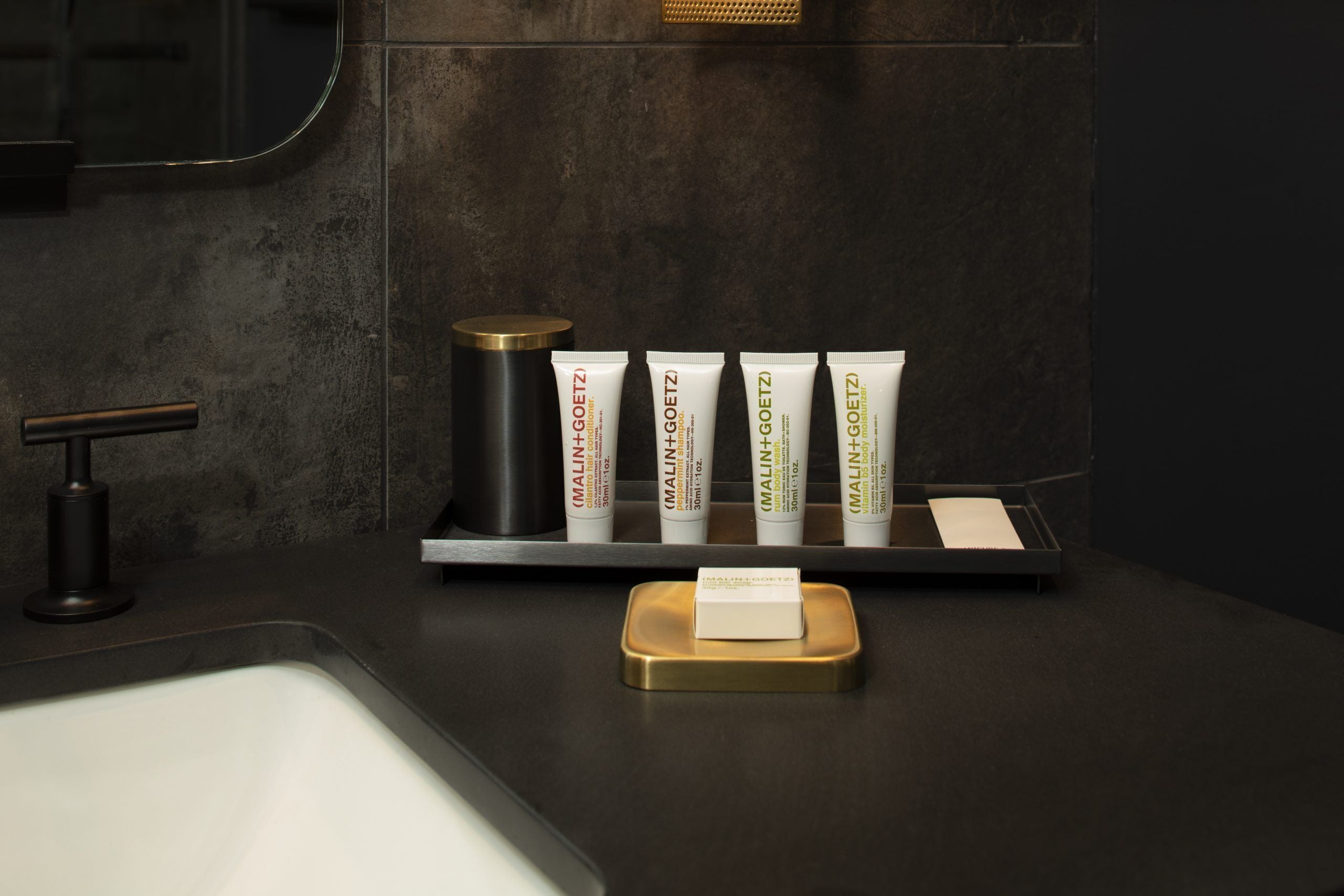 A bathroom counter with a black sink, a gold-colored soap dish, and five tubes of Malin+Goetz body lotion.