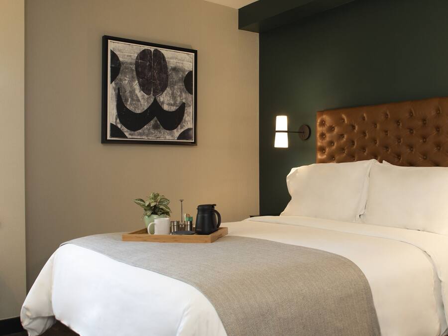A neatly made bed with a tufted headboard, a bedside table with a tray of items, a wall-mounted light, and a framed abstract painting.