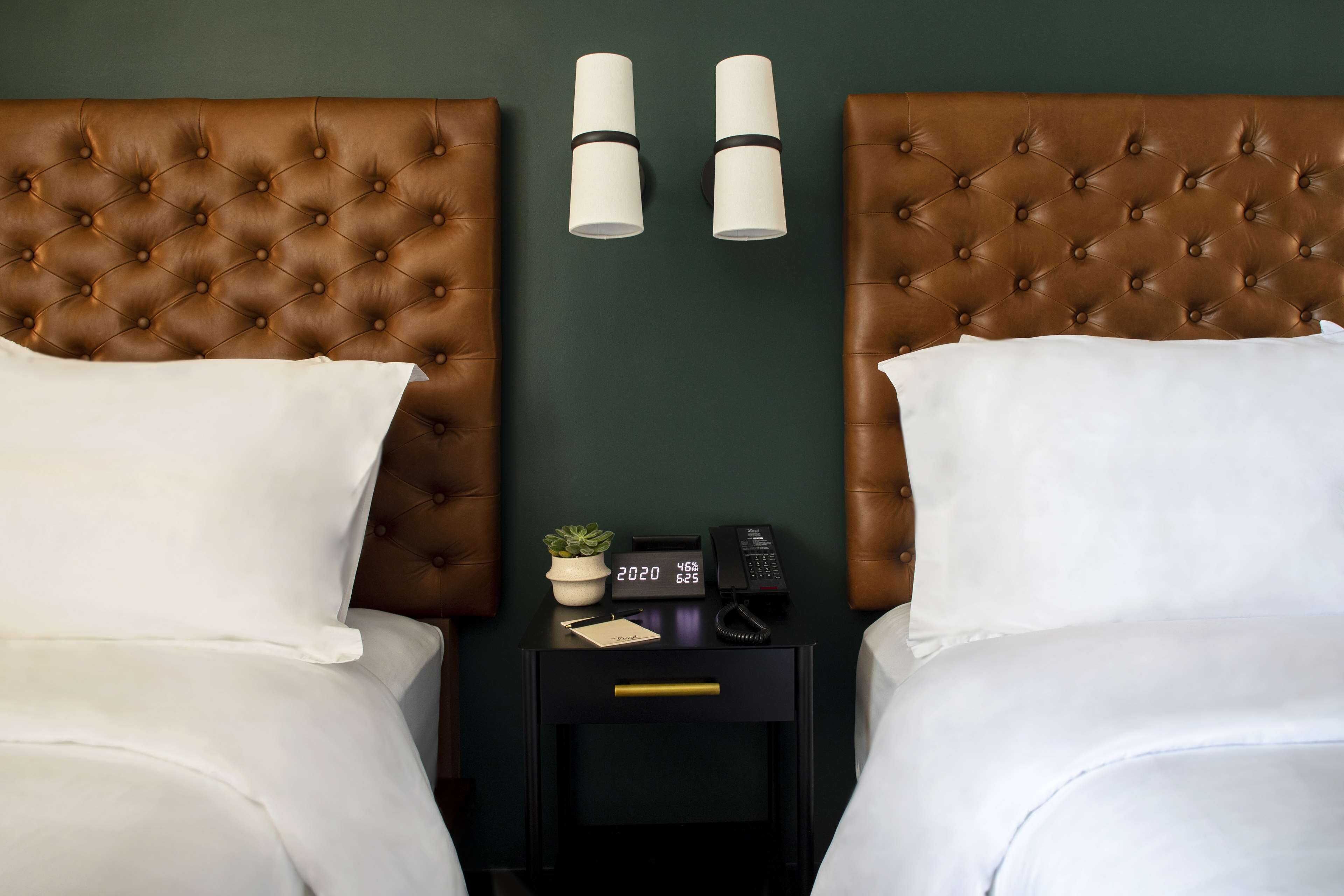 A neatly made twin bed with white linens, a small bedside table with a clock, a plant, and a phone.