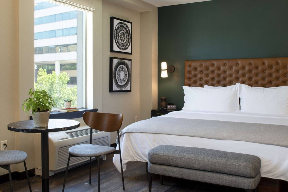 Une chambre d’hôtel avec un lit, une petite table sur laquelle est posée une plante en pot, une chaise et un banc. Il y a une fenêtre donnant sur les bâtiments à l’extérieur.