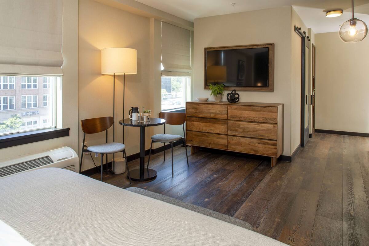 Une chambre d’hôtel comprenant un lit, un bureau avec deux chaises, une petite table, une lampe, une télévision et une commode.