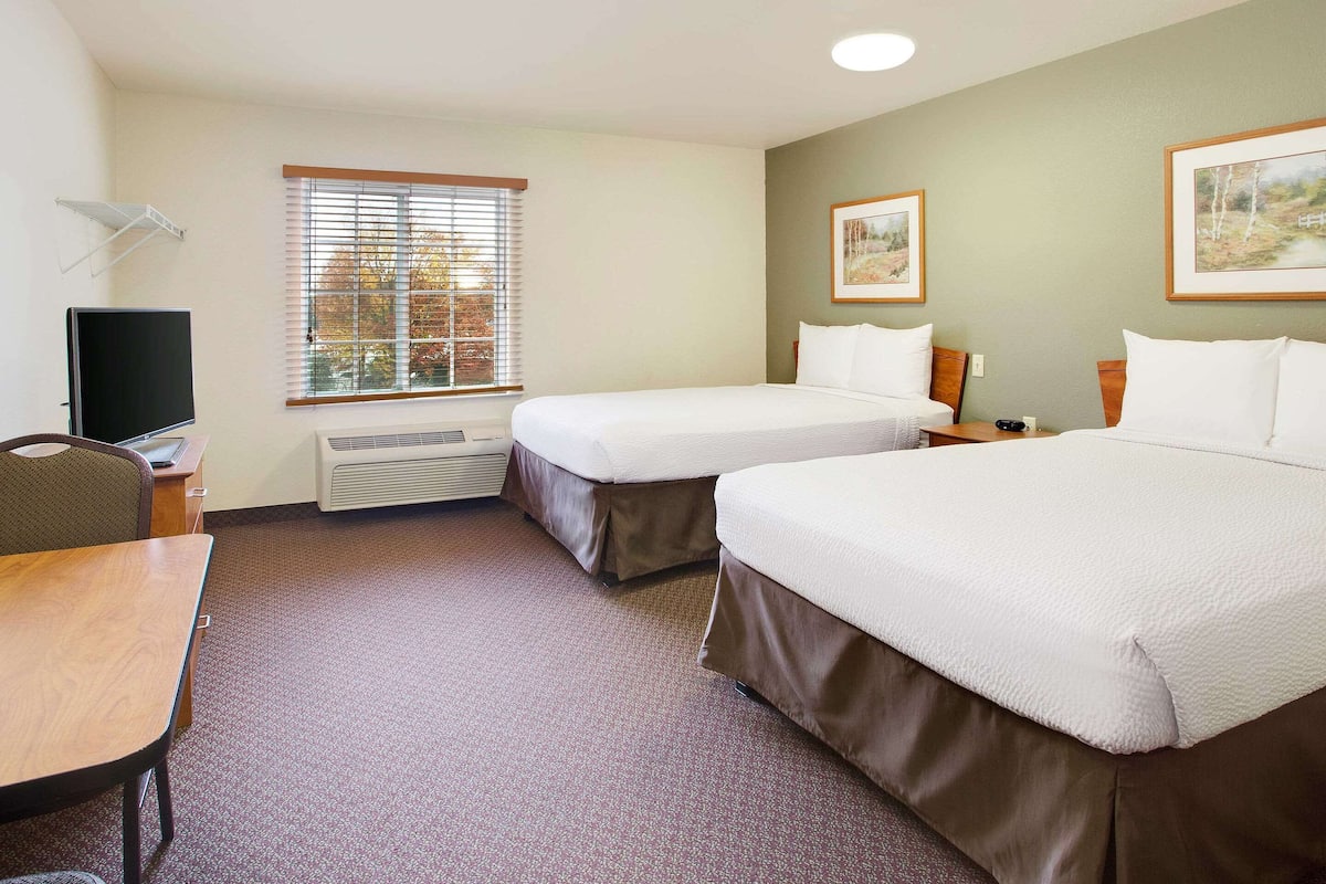 A hotel room with two beds, a desk, a chair, a TV, a window with blinds, and a radiator.