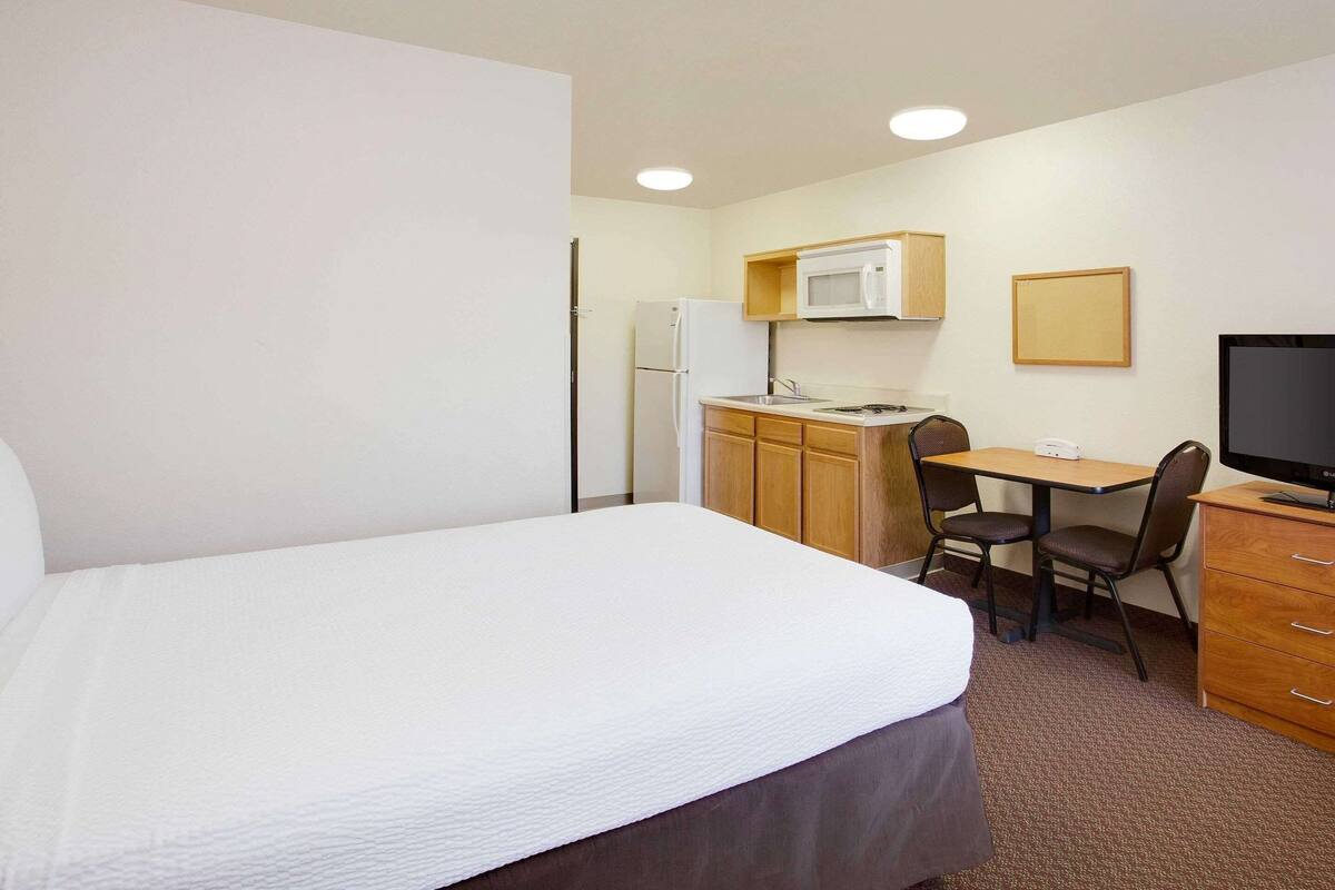 A hotel room with a bed, a desk with a chair, a microwave, a refrigerator, a television, and a wooden cabinet.