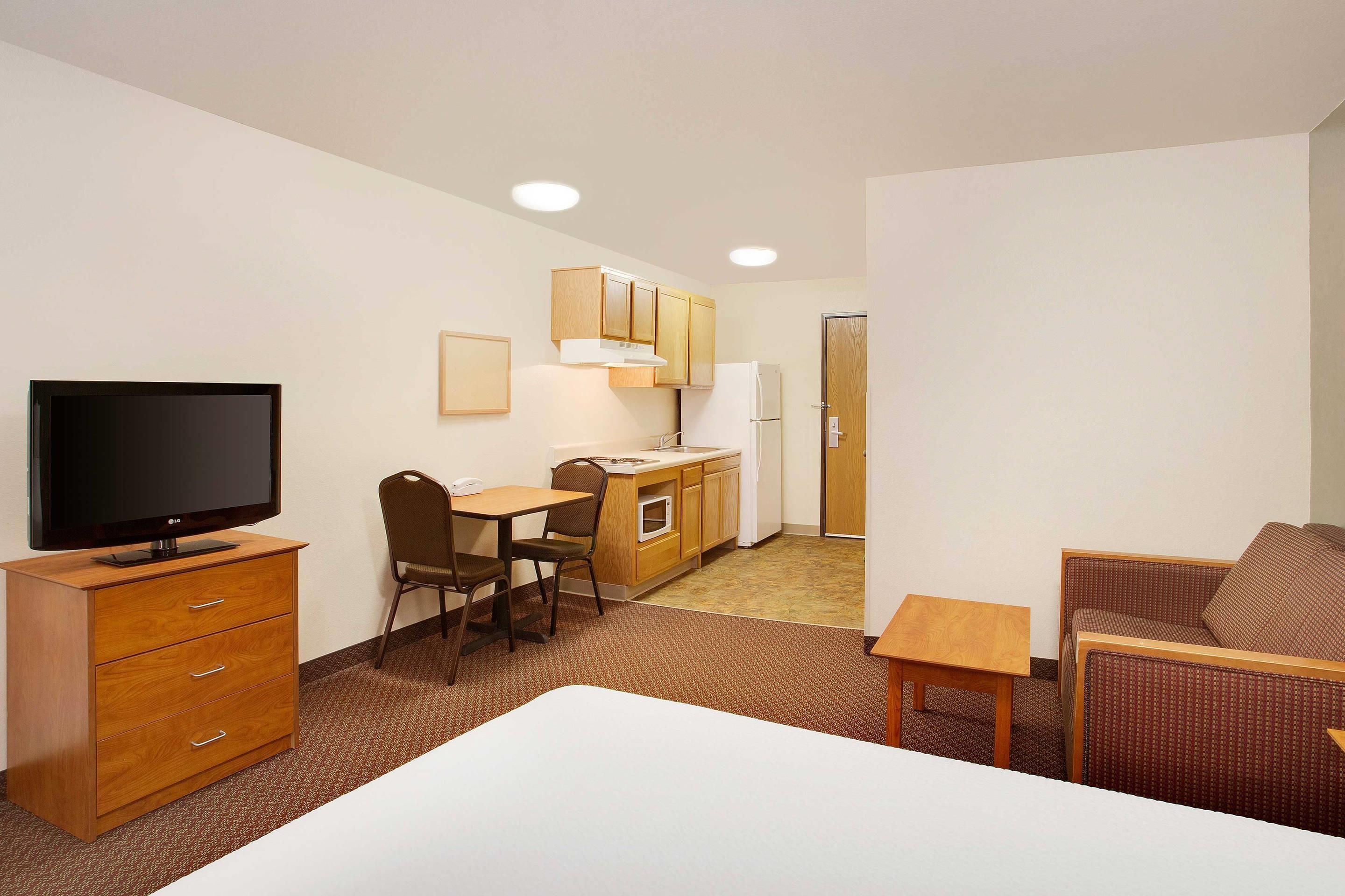 A compact hotel room with a television, a dining area, a kitchenette, and a seating area.