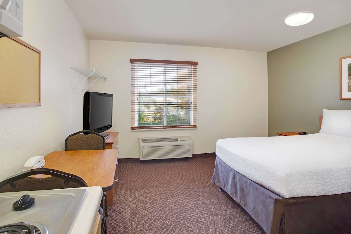 A hotel room with a bed, a desk, a chair, a TV, a window with blinds, and an air conditioning unit.