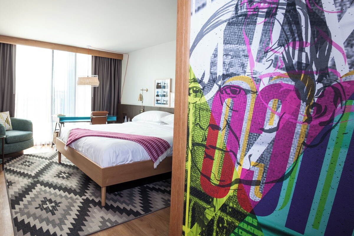 Une chambre d’hôtel avec un lit, un bureau, une chaise, une fenêtre avec des rideaux et un tableau au mur.