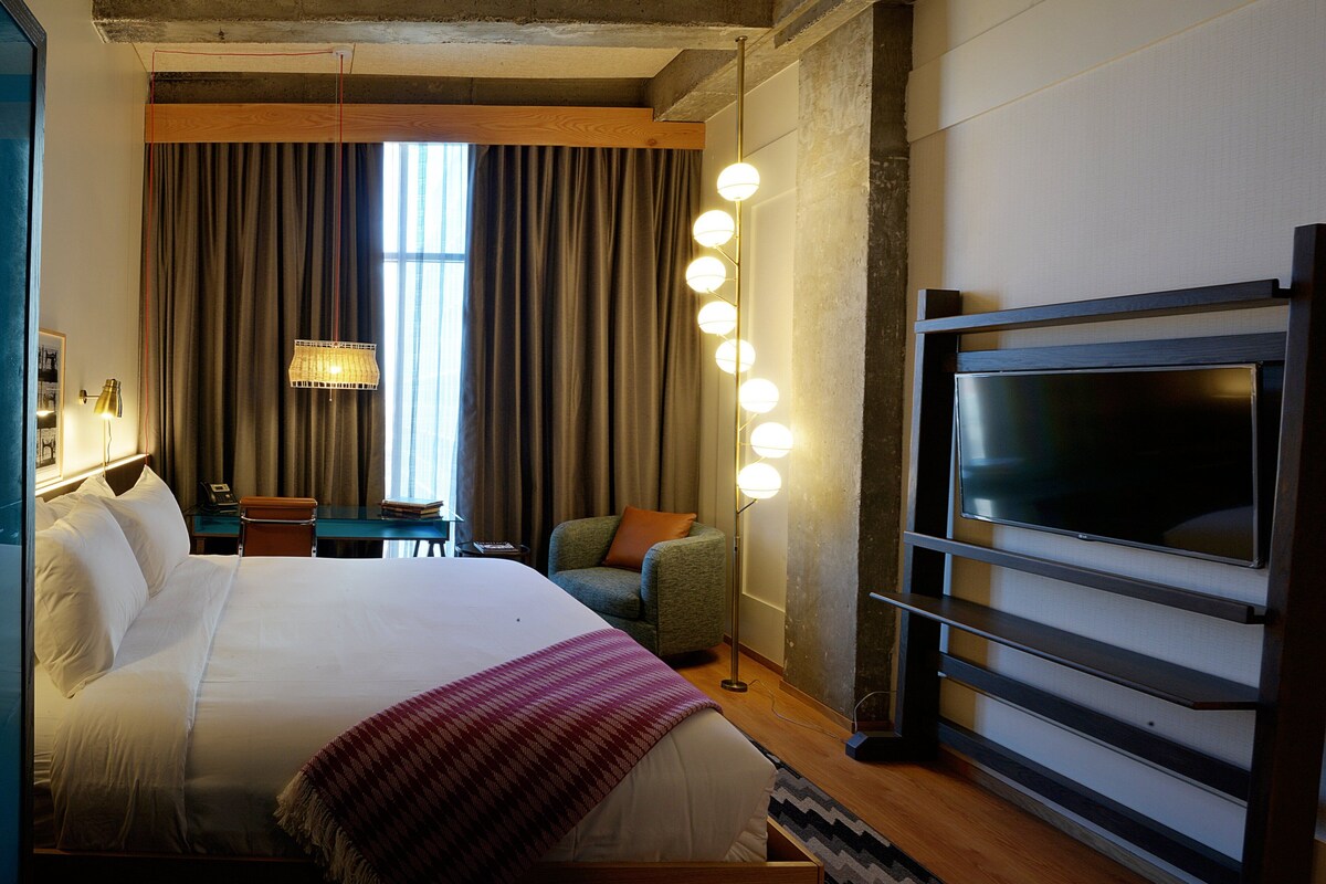 Une chambre d’hôtel avec un grand lit, un bureau avec une chaise, une télévision et une fenêtre avec des rideaux.