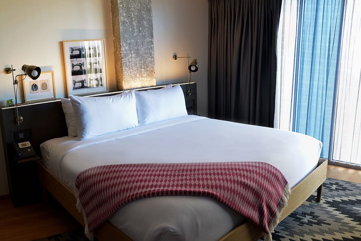 Une chambre d’hôtel avec un grand lit, une table de chevet avec une lampe, un tableau encadré au mur et une fenêtre avec des rideaux.