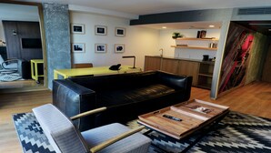 A modern living room with a black leather sofa, a wooden coffee table, and a yellow table. There is a kitchen area with wooden cabinets and a sink.