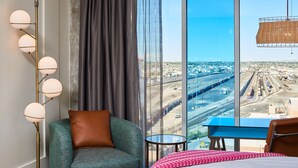 A hotel room with a large window, a sofa, a bed, a desk, and a floor lamp.