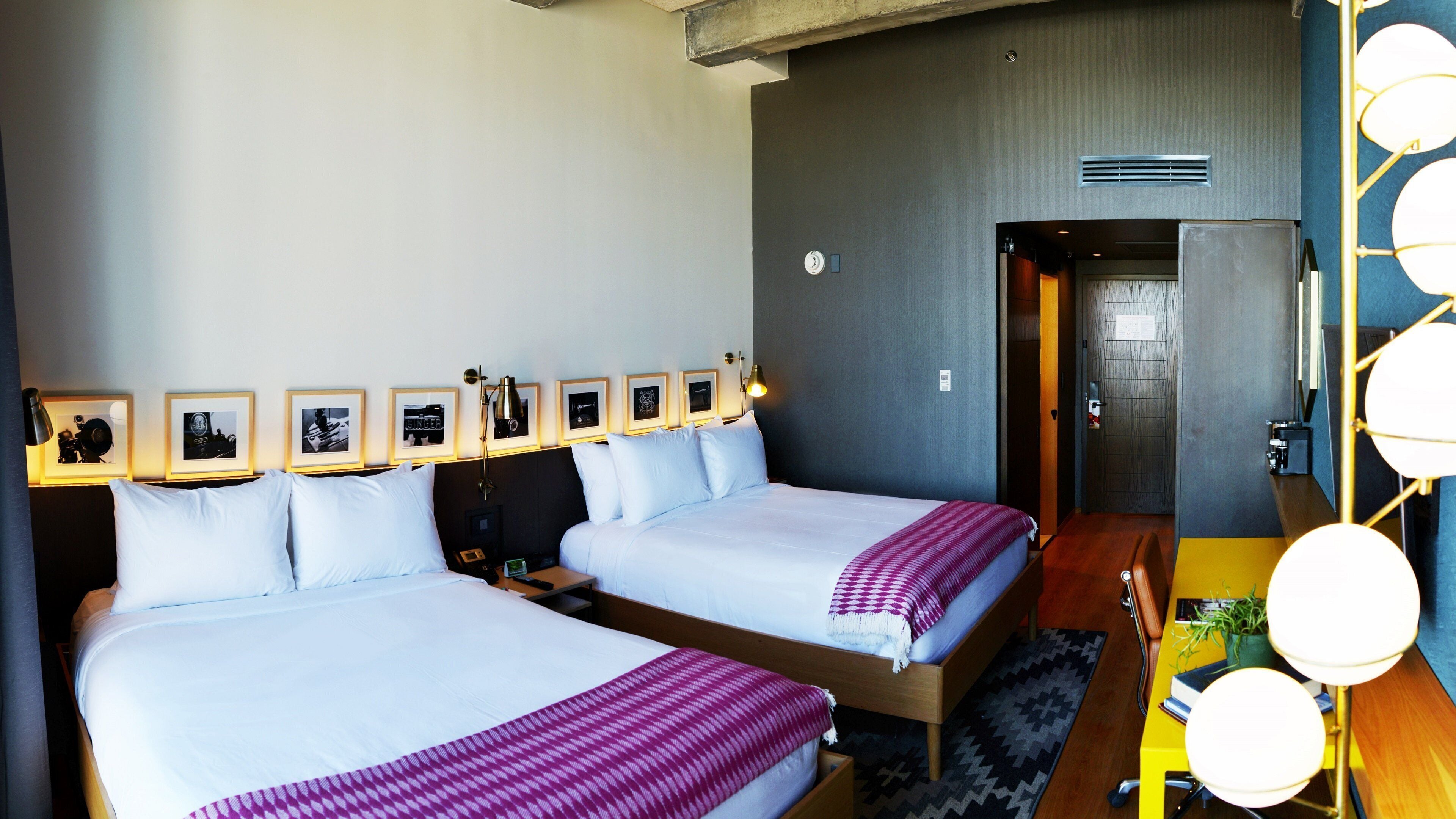 A hotel room with two beds, a desk, a chair, a lamp, framed pictures on the wall, and a door leading to another room.