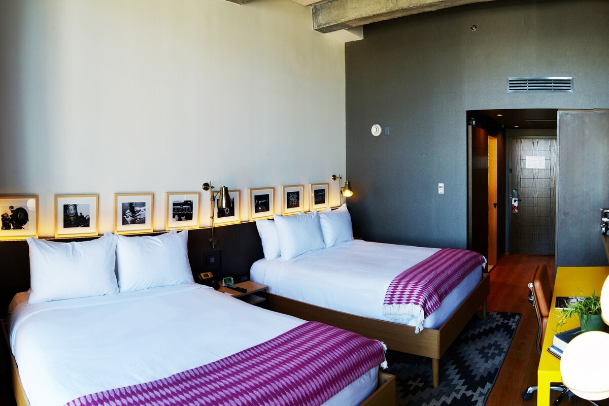 Une chambre d’hôtel avec deux lits, un bureau, une chaise, une lampe, des tableaux encadrés au mur et une porte donnant sur une autre pièce.