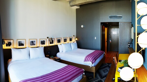 Habitación de hotel con dos camas, un escritorio, una silla, una lámpara, cuadros en la pared y una puerta que da a otra habitación.