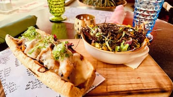 Un sándwich con queso y verduras sobre un papel con texto, un bol de ensalada y un vaso de agua sobre una mesa.