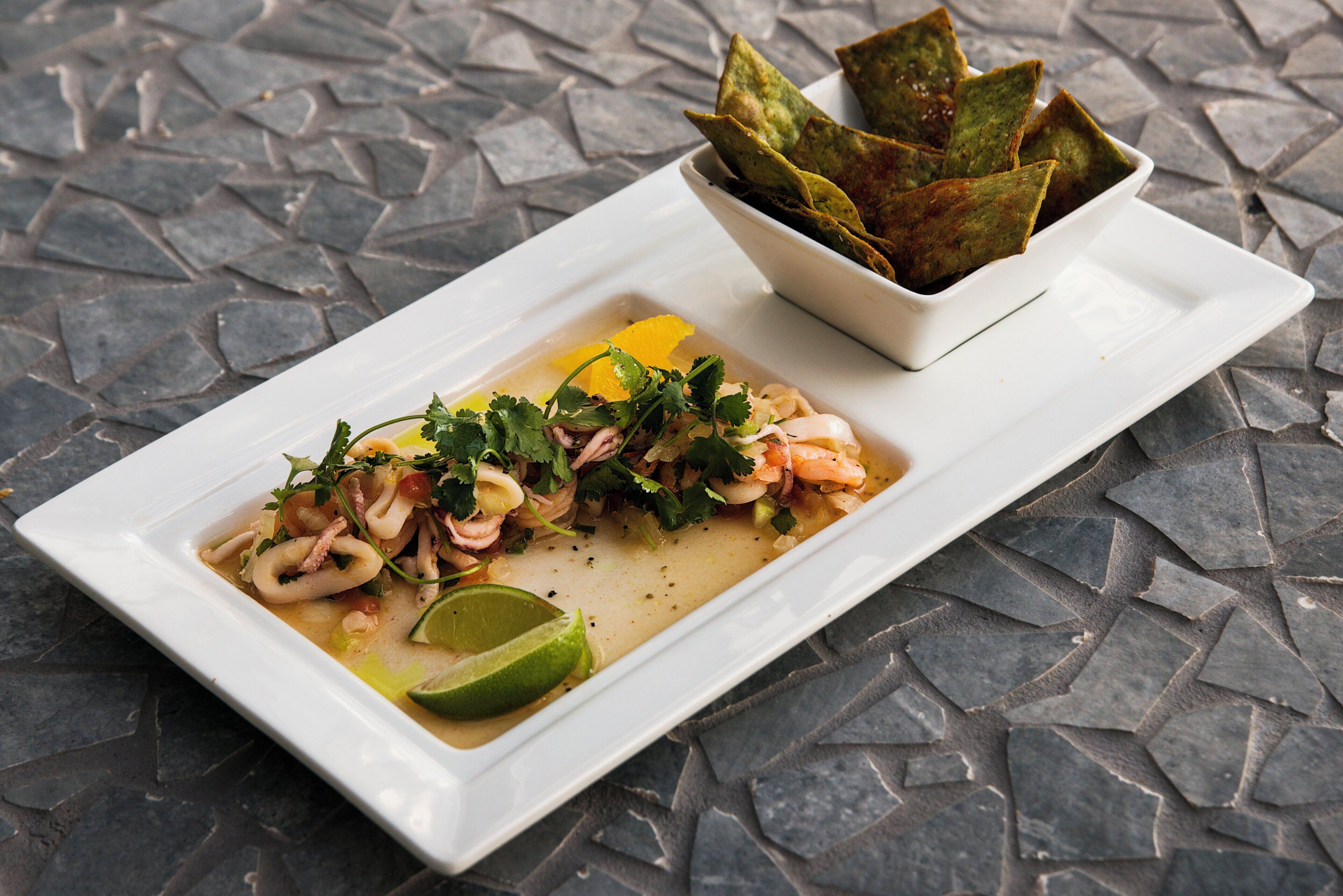 A rectangular white plate with a seafood dish, a lime wedge, and a bowl of green leaves on a stone-tiled surface.