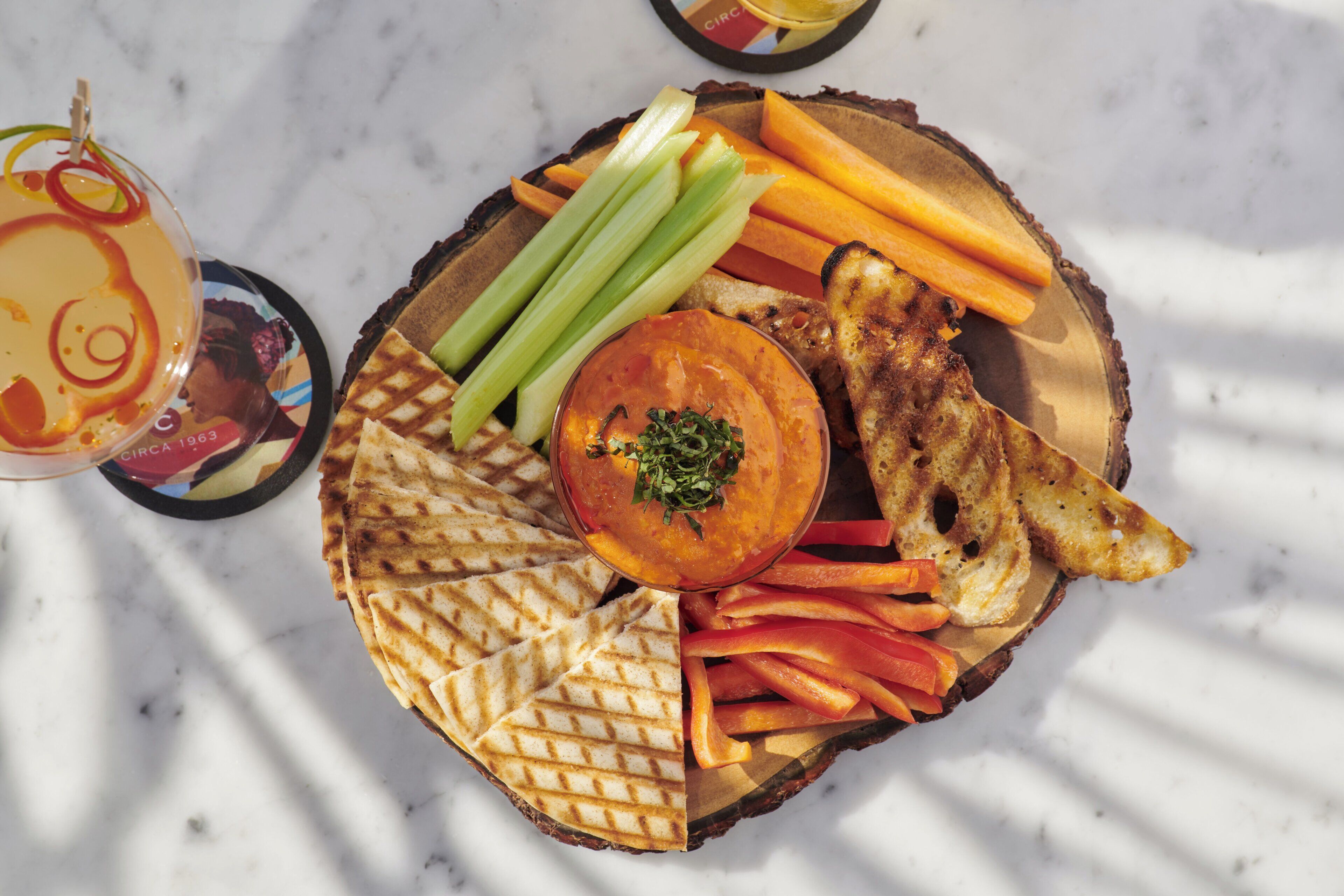 A wooden platter with a variety of foods including toast, carrots, celery, and a central dish, possibly a dip, on a marble surface.