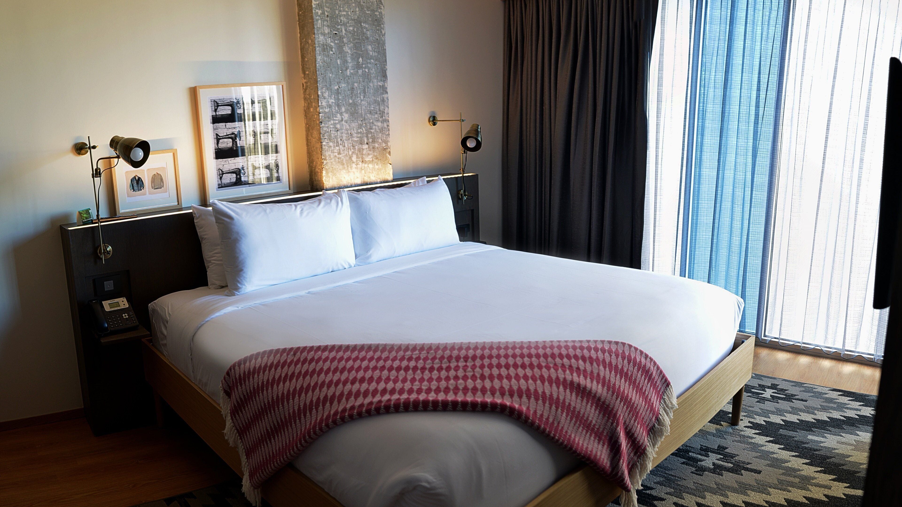 A hotel room with a large bed, a bedside table with a lamp, a framed picture on the wall, and a window with curtains.