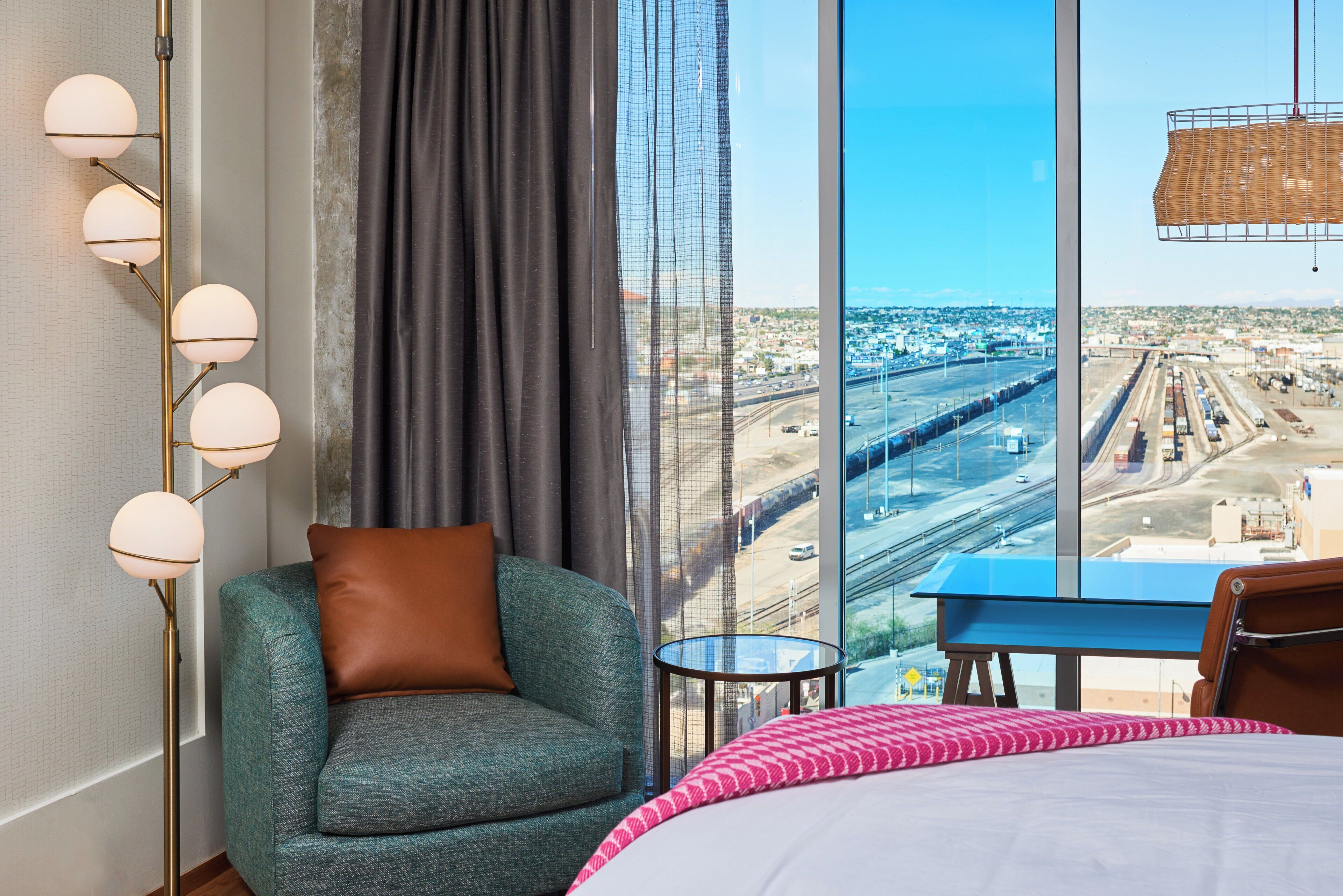 Une chambre d’hôtel avec une grande fenêtre, un lit, une chaise, une petite table et une lampe.
