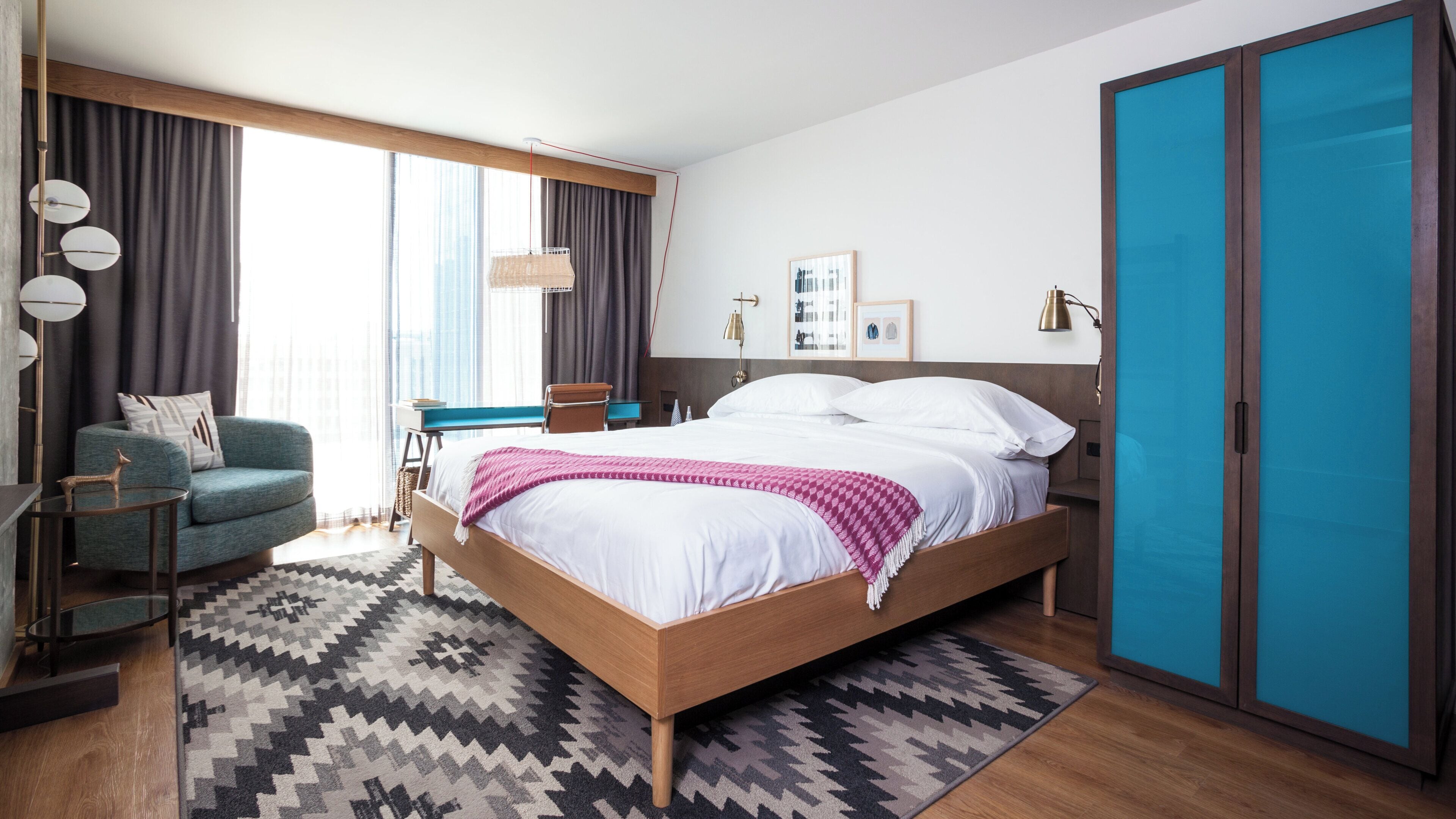 A modern hotel room with a bed, a desk, a chair, and a wardrobe.