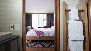 A hotel room with a bed, a wooden side table, a bench, a towel rack, and a window with curtains.
