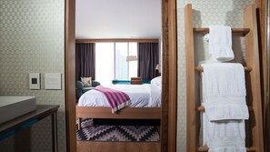 Une chambre d’hôtel comprenant un lit, une table de chevet en bois, un banc, un porte-serviettes et une fenêtre avec des rideaux.