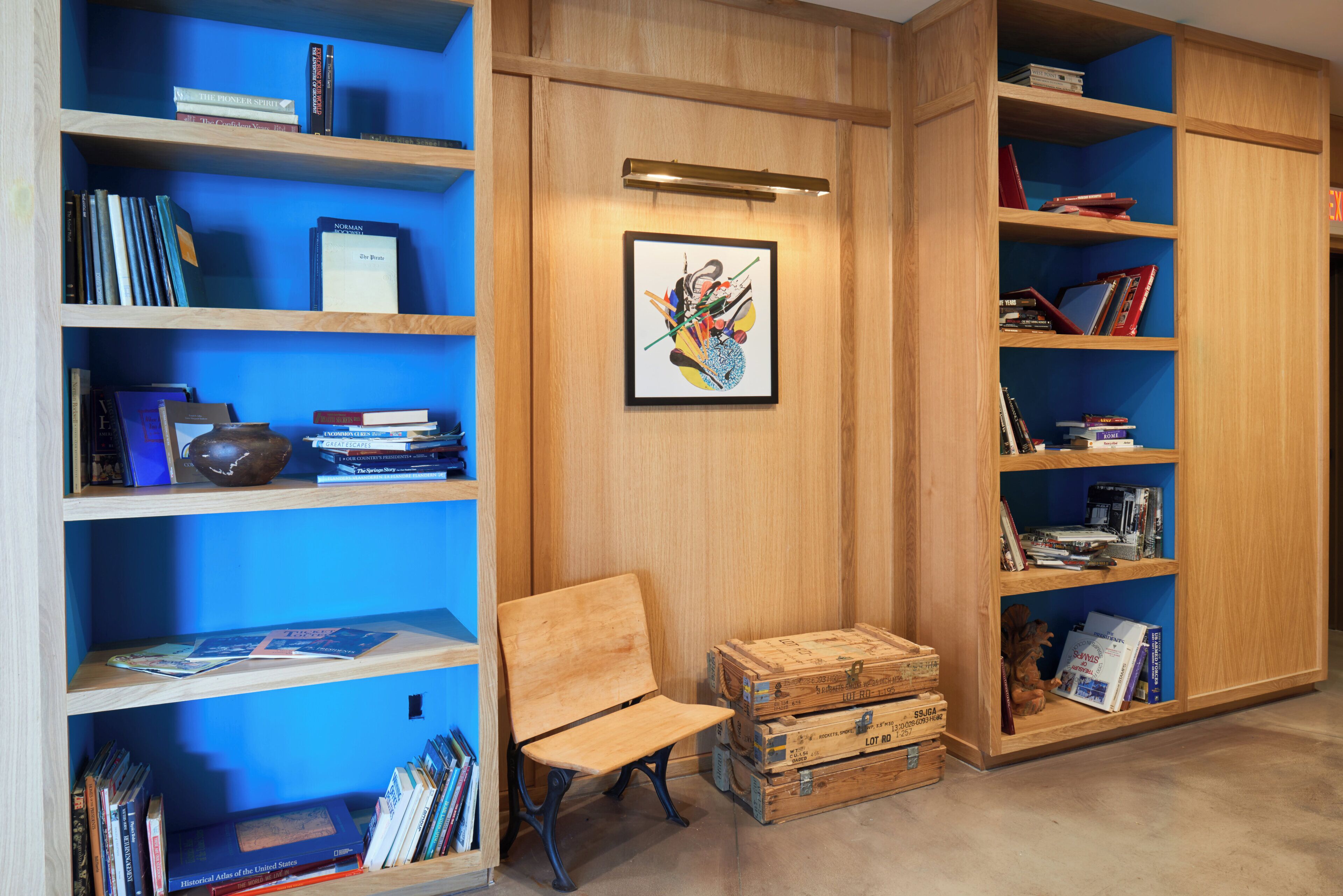 A library with wooden bookshelves, a framed abstract painting, a wooden bench, and vintage suitcases.
