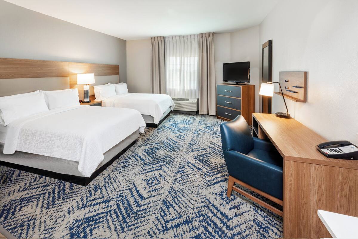 A hotel room with two beds, a desk, a chair, a TV, and a desk lamp.