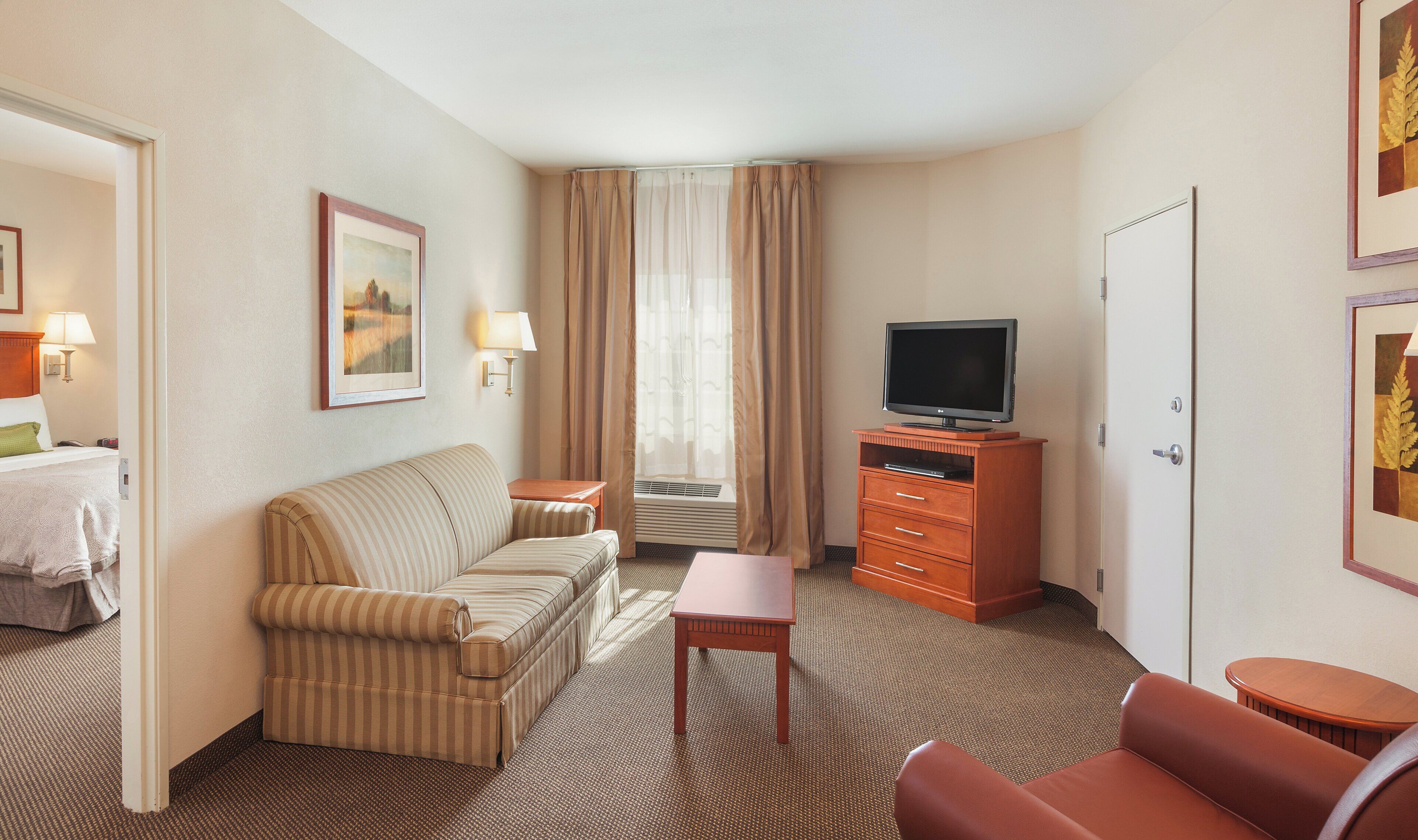 A hotel room with a striped sofa, a small table, a television on a wooden stand, a door, and a framed picture on the wall.