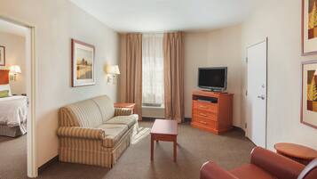 A hotel room with a striped sofa, a small table, a television on a wooden stand, a door, and a framed picture on the wall.
