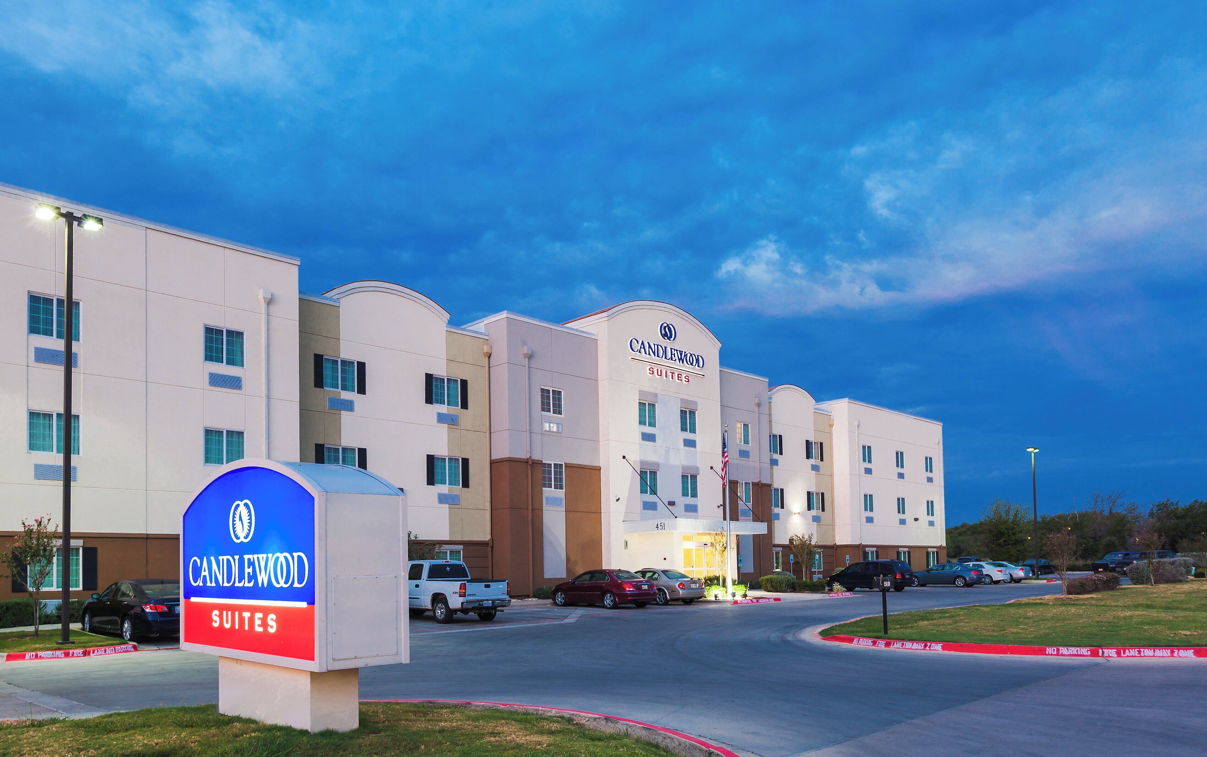 A hotel with a prominent sign for Candlewood Suites, multiple parking spaces, and a clear sky.