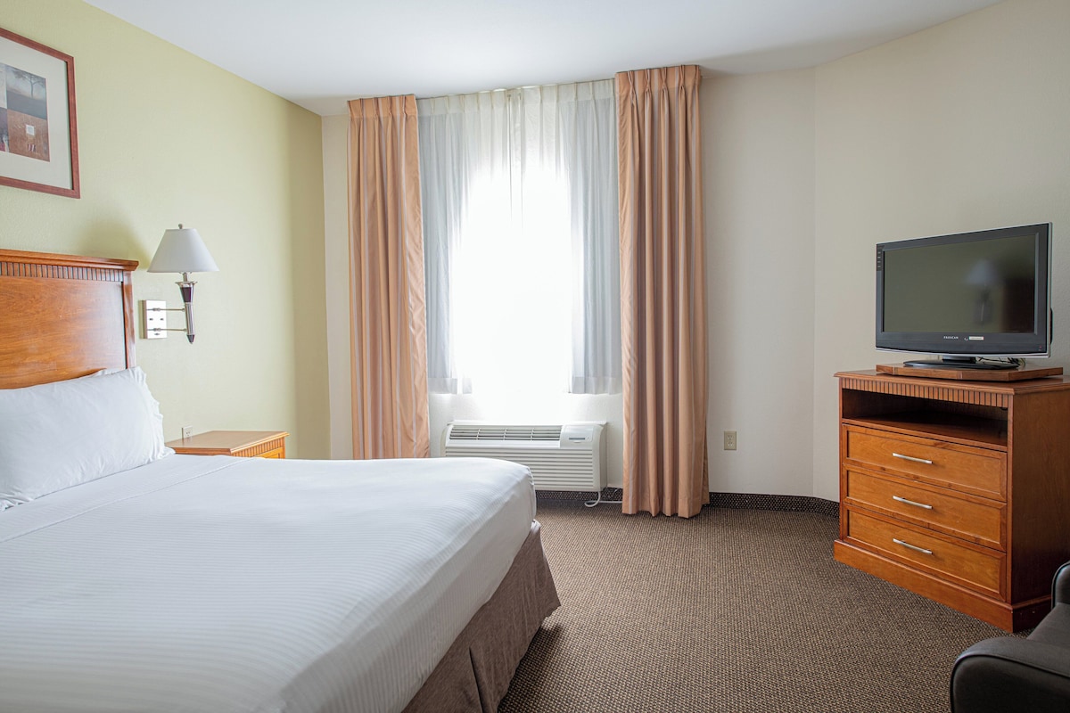 A hotel room with a bed, a television on a wooden stand, a desk with a lamp, and a window with curtains.