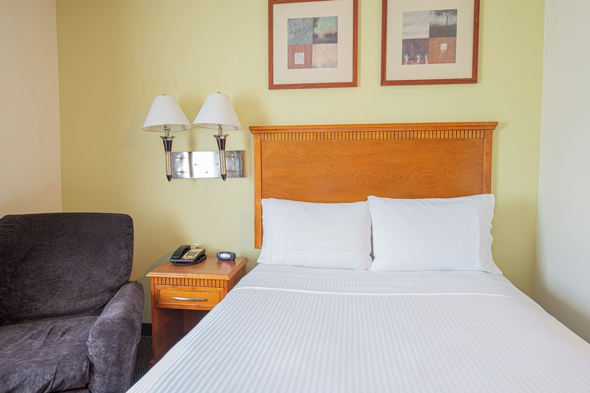 Quarto de hotel com uma cama grande, cabeceira de madeira, duas luminárias fixadas na parede, mesa de cabeceira com telefone e controle remoto, e uma poltrona cinza.
