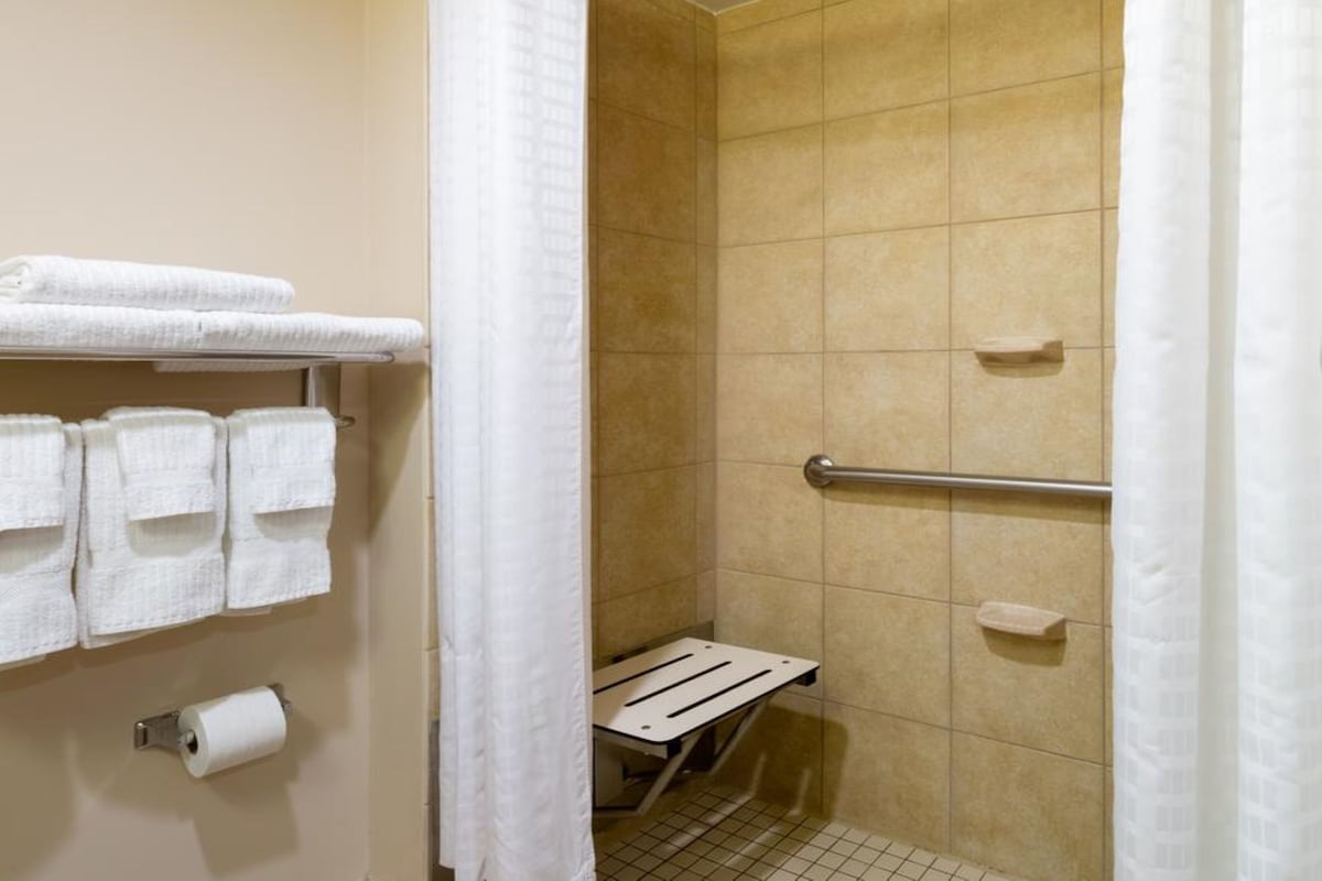 Pequeno espaço de chuveiro com azulejos, barra de apoio, porta-toalhas com toalhas dobradas e dispensador de papel higiénico.