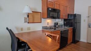 A compact kitchen with wooden cabinets, a microwave, a refrigerator, a coffee maker, and a dining table with chairs. - Candlewood Suites New Iberia by IHG (New Iberia)