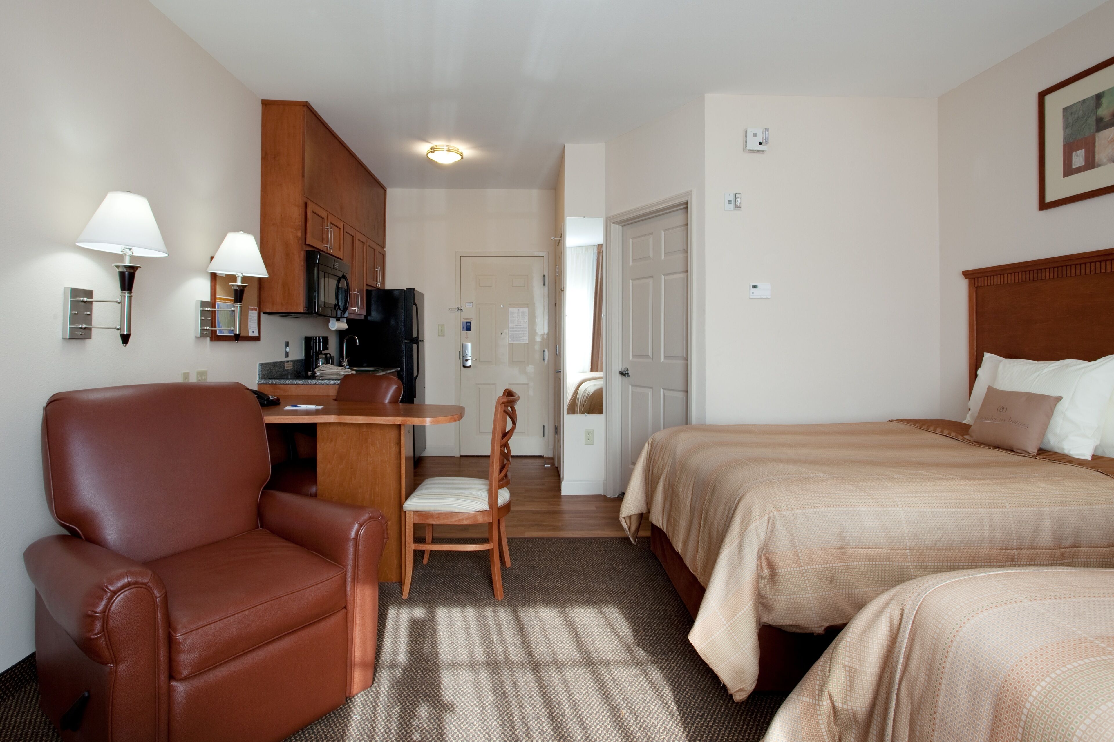 Habitación de hotel con cama, escritorio, silla, kitchenette y una puerta que da a otra habitación.
