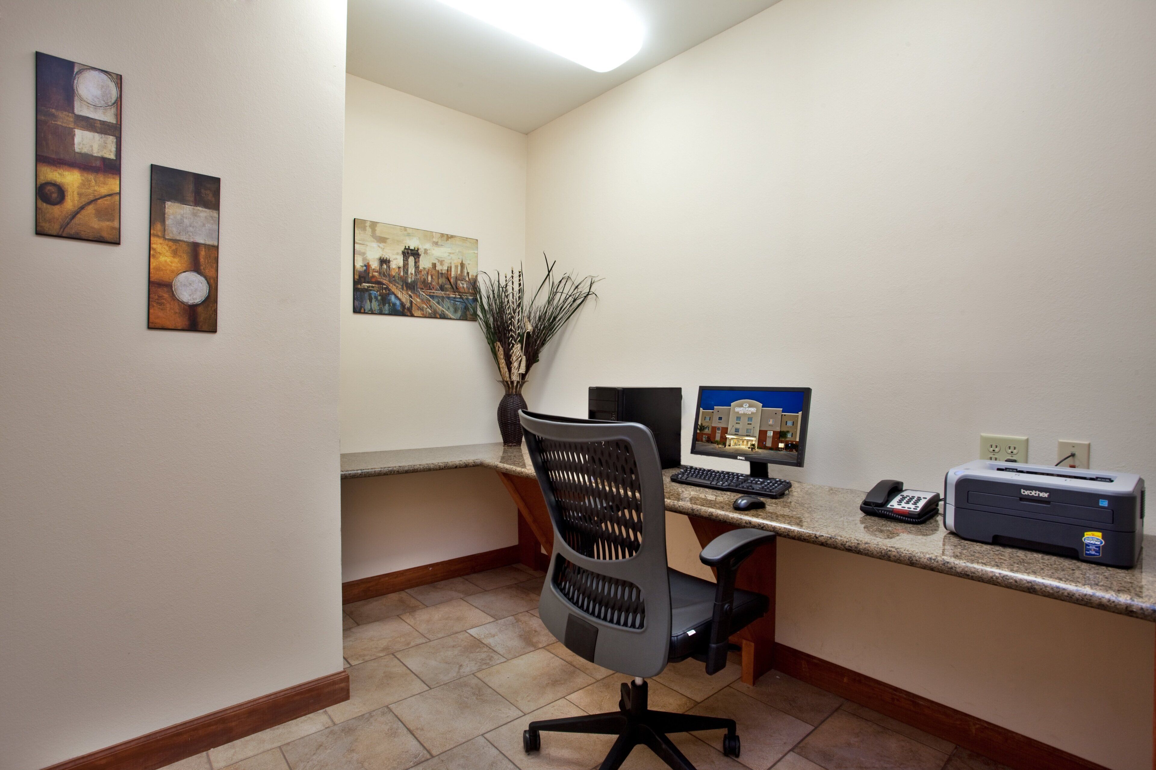 A small office with a desk, computer, printer, and a chair. There are two paintings on the wall and a vase with plants on the desk.